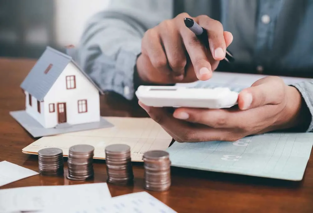 A person sitting at a desk with a calculator, a tiny house, bank books, and stacks of quarters.