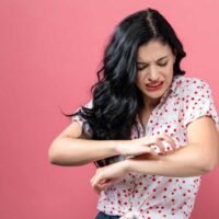 Woman standing in front of a pink background scratching her arm