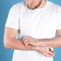 Man wearing a white shirt against a blue background scratching one arm with the opposite hand.