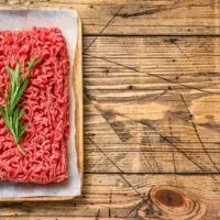 Raw ground beef on a wooden table topped with a sprig of rosemary